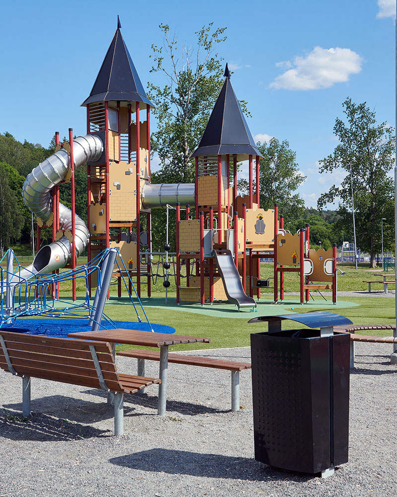 Al fondo se ve una gran torre de juegos, con un banco de parque y una papelera en primer plano para que los padres puedan tener visibilidad de los niños mientras se relajan.